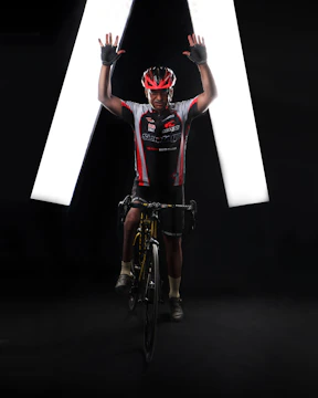 A cyclist wearing a red and white cycling jersey and helmet poses on a bicycle with both arms raised. The background is dark with bright white lights forming an inverted V shape behind the cyclist.