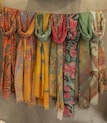 A row of vibrant, patterned scarves hang on a line against a textured wall. The scarves display intricate designs in various colors, including floral and paisley patterns. They range in hues from warm yellows and oranges to cool blues and greens.