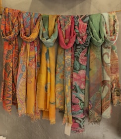 A row of vibrant, patterned scarves hang on a line against a textured wall. The scarves display intricate designs in various colors, including floral and paisley patterns. They range in hues from warm yellows and oranges to cool blues and greens.