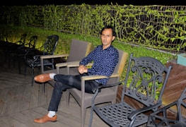A person is seated outdoors in a relaxed posture. The setting includes metal and woven chairs, with a wooden deck floor. A lush green vine-covered fence serves as a backdrop, and there are decorative chairs around.