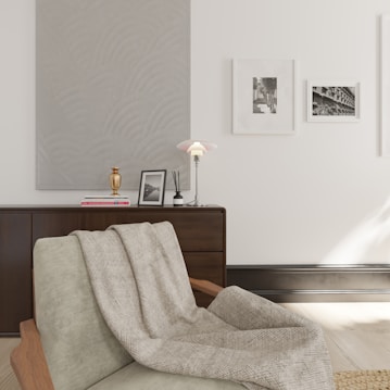 A cozy interior setting with a sleek armchair draped with a textured grey blanket. A modern lamp with a soft glow sits on a wooden sideboard alongside framed black-and-white photographs and a decorative golden vase. The wall features minimalist art and is painted in a soft, neutral tone.