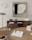 A modern interior scene featuring a wooden dining table with a woven rattan chair and a round clock on the wall. A sleek, minimalistic art piece hangs above a wooden sideboard, which holds a digital clock, camera, and a gaming console. Books are stacked on the sideboard and table, and a small plant adds a touch of greenery.