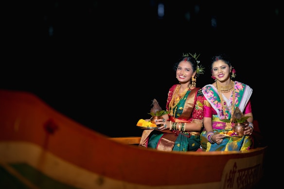 Two women dressed in traditional, colorful Indian attire and jewelry are sitting in a boat holding decorated items. They are smiling against a dark background, suggesting a festive or cultural setting.