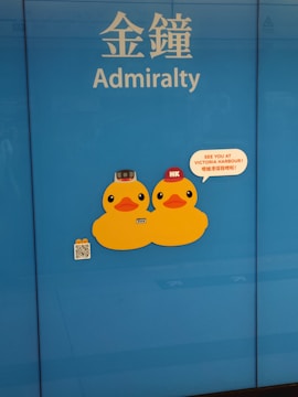 Two cartoon duck stickers are displayed on a blue background. Above them are the words 'Admiralty' in English and Chinese. One duck is wearing a brown helmet and the other a red cap with 'HK' on it. To the right, a speech bubble says 'See you at Victoria Harbour!'. A small QR code and additional small duck images are visible below the ducks.