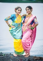 Two women dressed in vibrant traditional attire stand on a rocky surface by the water. Both wear ornate sarees in contrasting colors of yellow and pink, complemented by intricate gold jewelry. Their hair is adorned with flowers, enhancing the festive appearance. They pose playfully, exuding a sense of joy and celebration.