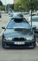 A grey BMW car is parked in a lot with a roof cargo box on top. The windshield has a large decal reading 'MISA1TED'. The license plate reads 'JA-5359'. A tree is visible next to the car, and other vehicles are seen in the background under a cloudy sky.
