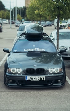 A grey BMW car is parked in a lot with a roof cargo box on top. The windshield has a large decal reading 'MISA1TED'. The license plate reads 'JA-5359'. A tree is visible next to the car, and other vehicles are seen in the background under a cloudy sky.