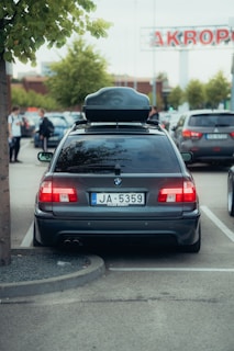 A parked black BMW station wagon with a roof box on top, located in a parking lot. The license plate is visible, with the prefix 'JA' and series of numbers. In the background, there are other cars and a shopping center sign partially visible, along with a few trees lining the area and several people walking.