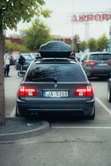 A parked black BMW station wagon with a roof box on top, located in a parking lot. The license plate is visible, with the prefix 'JA' and series of numbers. In the background, there are other cars and a shopping center sign partially visible, along with a few trees lining the area and several people walking.