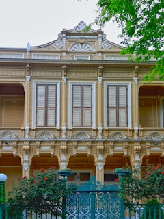 An intricately designed wooden facade of a two-story building features ornate carvings and arched windows. The closed wooden shutters add a sense of uniformity, while vibrant red flowers and lush greenery accentuate the decorative iron gate in the foreground. The architectural style is influenced by historical design elements, showcasing an elegant and timeless aesthetic.