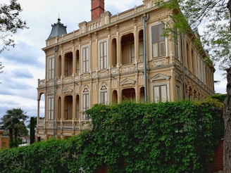 A large, ornate, beige mansion with intricate architectural details, including columns and decorative moldings. The building has multiple windows with shutters and a prominent chimney. Lush green foliage partially covers the lower part of the mansion. The sky is cloudy, adding a dramatic backdrop.