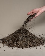 A hand is pouring a pile of gravelly soil onto a beige surface. The soil is a mixture of variously sized pebbles and dirt, forming a small mound.