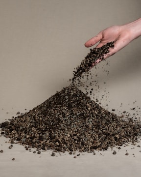 A hand is pouring a pile of gravelly soil onto a beige surface. The soil is a mixture of variously sized pebbles and dirt, forming a small mound.