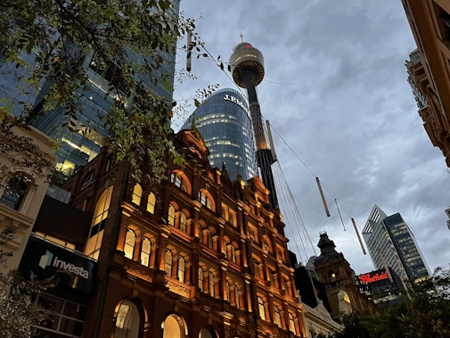 A bustling urban scene with modern skyscrapers and a historic red brick building illuminated by warm lights. Above, a prominent tower rises against a backdrop of a cloudy evening sky. Signs for 'investa' and 'Westfield' are visible among the buildings. Leafy branches partially frame the view from the left.