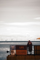 A lifeguard station is positioned by a large body of water with wind turbines visible in the distance across the horizon. A person dressed in lifeguard attire leans on the railing, overseeing the water. The atmosphere appears tranquil with calm water and a cloudy sky.