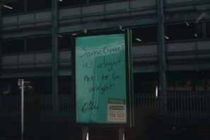 A green signboard features handwritten text that reads 'Sometimes it's alright not to be alright.' Below is information about a helpline, Samaritans, with a contact number and website. The background includes a dark, industrial-looking building with multiple floors and a metal fence.