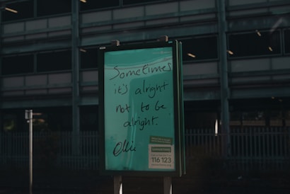 A green signboard features handwritten text that reads 'Sometimes it's alright not to be alright.' Below is information about a helpline, Samaritans, with a contact number and website. The background includes a dark, industrial-looking building with multiple floors and a metal fence.