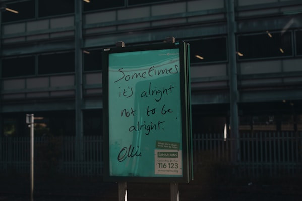 A green signboard features handwritten text that reads 'Sometimes it's alright not to be alright.' Below is information about a helpline, Samaritans, with a contact number and website. The background includes a dark, industrial-looking building with multiple floors and a metal fence.