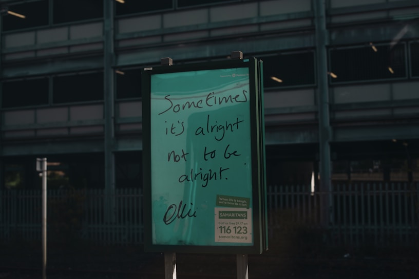A green signboard features handwritten text that reads 'Sometimes it's alright not to be alright.' Below is information about a helpline, Samaritans, with a contact number and website. The background includes a dark, industrial-looking building with multiple floors and a metal fence.