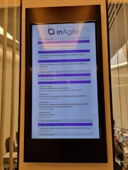 A digital display in a modern setting shows an agenda for a conference or workshop related to agile product development. The schedule includes various sessions, case studies, and coffee breaks, with timings from 9:00 to 16:45. The display has a header labeled 'Product inAgile' and sections are highlighted in purple.