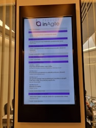 A digital display in a modern setting shows an agenda for a conference or workshop related to agile product development. The schedule includes various sessions, case studies, and coffee breaks, with timings from 9:00 to 16:45. The display has a header labeled 'Product inAgile' and sections are highlighted in purple.
