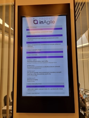 A digital display in a modern setting shows an agenda for a conference or workshop related to agile product development. The schedule includes various sessions, case studies, and coffee breaks, with timings from 9:00 to 16:45. The display has a header labeled 'Product inAgile' and sections are highlighted in purple.