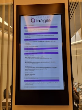 A digital display in a modern setting shows an agenda for a conference or workshop related to agile product development. The schedule includes various sessions, case studies, and coffee breaks, with timings from 9:00 to 16:45. The display has a header labeled 'Product inAgile' and sections are highlighted in purple.