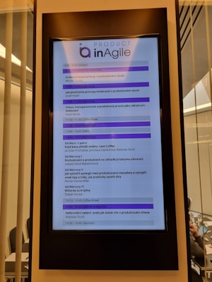A digital display in a modern setting shows an agenda for a conference or workshop related to agile product development. The schedule includes various sessions, case studies, and coffee breaks, with timings from 9:00 to 16:45. The display has a header labeled 'Product inAgile' and sections are highlighted in purple.