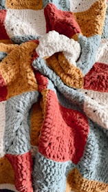A textured, multi-colored knitted blanket with thick yarn in shades of red, yellow, blue-gray, and white, forming a patchwork pattern.