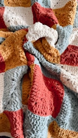 A textured, multi-colored knitted blanket with thick yarn in shades of red, yellow, blue-gray, and white, forming a patchwork pattern.