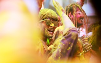 A group of people is joyfully celebrating, covered in vibrant colored powders. They appear to be participating in a festival or event similar to Holi, with laughter and energy evident on their faces. The crowd is tightly packed, and there's a sense of movement and excitement, with arms raised in the air.