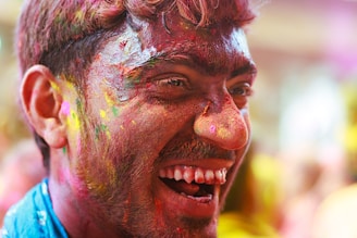 A close-up of a person with a joyful expression, smiling widely. Their face is covered in vibrant multicolored powders, suggesting a celebration or festival involving colorful substances. The background is soft-focused, adding to the lively atmosphere.