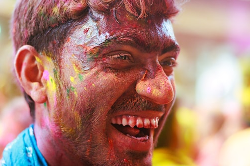 A close-up of a person with a joyful expression, smiling widely. Their face is covered in vibrant multicolored powders, suggesting a celebration or festival involving colorful substances. The background is soft-focused, adding to the lively atmosphere.