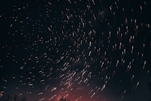 Long exposure photograph of the night sky capturing star trails in circular patterns across the dark canvas. The bottom of the image softly glows with a reddish hue, hinting at the horizon or distant light pollution. Sparse silhouettes of tree branches frame the lower corners.