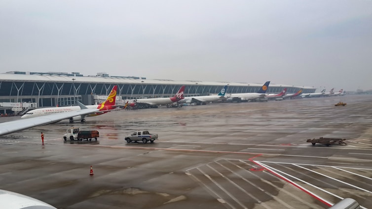Several airplanes are parked at a large airport terminal with a modern architectural design. The wet runway reflects the cloudy sky, indicating recent rain. Ground service vehicles are present near the planes, and a few airport staff are visible on the tarmac.