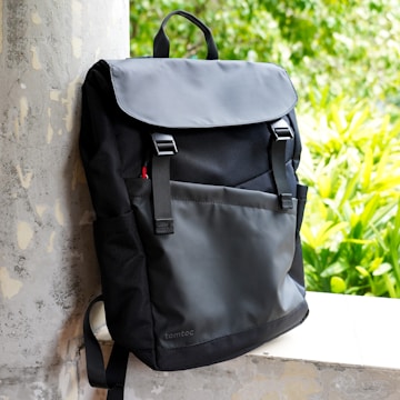 A sleek black backpack with a minimalist design leans against a textured concrete pillar. The scene is brightened by lush green foliage in the background, adding a natural contrast to the modern style of the bag.