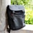 A sleek black backpack with a minimalist design leans against a textured concrete pillar. The scene is brightened by lush green foliage in the background, adding a natural contrast to the modern style of the bag.