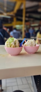 Two bowls of shaved ice dessert with a topping, possibly green jelly or syrup, placed on a light-colored tabletop. Each bowl is pink with a small spoon, one blue and one red. The background suggests a busy indoor area with people, but they are out of focus.