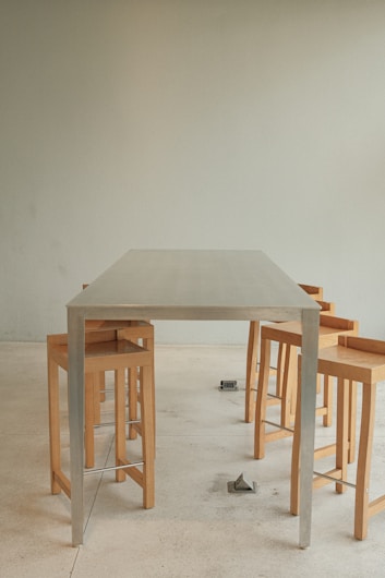 A modern dining setup with a minimalist metal table and several wooden stools arranged around it. The setting is simple with a neutral-toned wall and a light-colored floor, creating a clean and contemporary look.