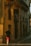 A person wearing a black shirt and red shorts walks down a narrow street lined with buildings. The warm sunlight casts long shadows on the walls. A sign with the text 'PÁLOMAR GROUP' is prominently visible on one of the buildings with a yellow facade.