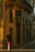 A person wearing a black shirt and red shorts walks down a narrow street lined with buildings. The warm sunlight casts long shadows on the walls. A sign with the text 'PÁLOMAR GROUP' is prominently visible on one of the buildings with a yellow facade.