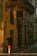 A person wearing a black shirt and red shorts walks down a narrow street lined with buildings. The warm sunlight casts long shadows on the walls. A sign with the text 'PÁLOMAR GROUP' is prominently visible on one of the buildings with a yellow facade.