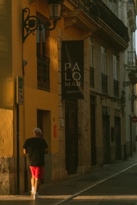 A person wearing a black shirt and red shorts walks down a narrow street lined with buildings. The warm sunlight casts long shadows on the walls. A sign with the text 'PÁLOMAR GROUP' is prominently visible on one of the buildings with a yellow facade.