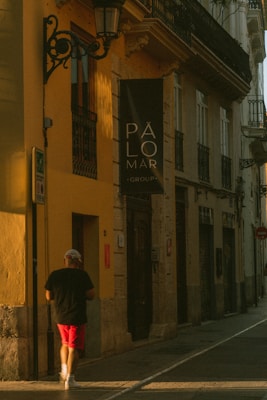 A person wearing a black shirt and red shorts walks down a narrow street lined with buildings. The warm sunlight casts long shadows on the walls. A sign with the text 'PÁLOMAR GROUP' is prominently visible on one of the buildings with a yellow facade.