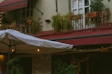 A cozy outdoor setting featuring a building with potted plants on the window sills above, and large red awnings providing shelter. Below, a large umbrella with string lights is partially visible, casting a warm glow and enhancing the rustic ambiance.