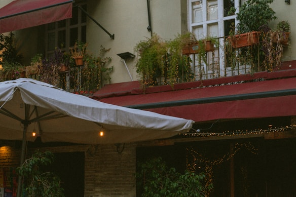 A cozy outdoor setting featuring a building with potted plants on the window sills above, and large red awnings providing shelter. Below, a large umbrella with string lights is partially visible, casting a warm glow and enhancing the rustic ambiance.