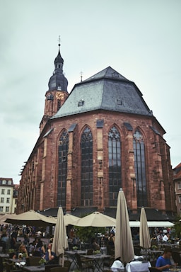 A large, historic church with a tall steeple and intricate Gothic architecture, featuring pointed arches and tall, narrow windows. In front of the church, a bustling outdoor café scene is present, with people seated under tan umbrellas, enjoying their time.