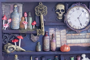 A collection of vintage apothecary bottles, ornate keys, and small red and white mushrooms are arranged on shelves. There is an antiquated clock with Roman numerals, a decorative skull, and a miniature pumpkin nearby, all set against a backdrop of old books.