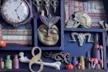 A shelf filled with various curiosities, including a vintage clock with Roman numerals, small decorative skulls, an ornate face mask, and tiny bottles. The shelves are lined with fake books, and there are small mushrooms and bone replicas adding a mystical feel.