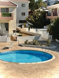 A serene residential area featuring a small, clear blue swimming pool surrounded by a tiled patio. Adjacent to the pool is a white reclining lounge chair. In the background, several two-story houses with red-tiled roofs and plant-lined balconies can be seen. There is also a small set of outdoor furniture, including a table and chairs, and various green plants and trees surround the area.